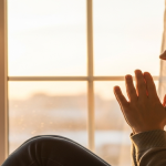 Mujer sentada en un alféizar de ventana, con las manos juntas en actitud de oración.